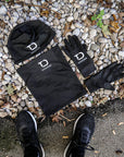 Lieferumfang des schwarzen Tapedesign Wintersets: Beanie, Halswärmer und ein Paar Handschuhe auf Kies