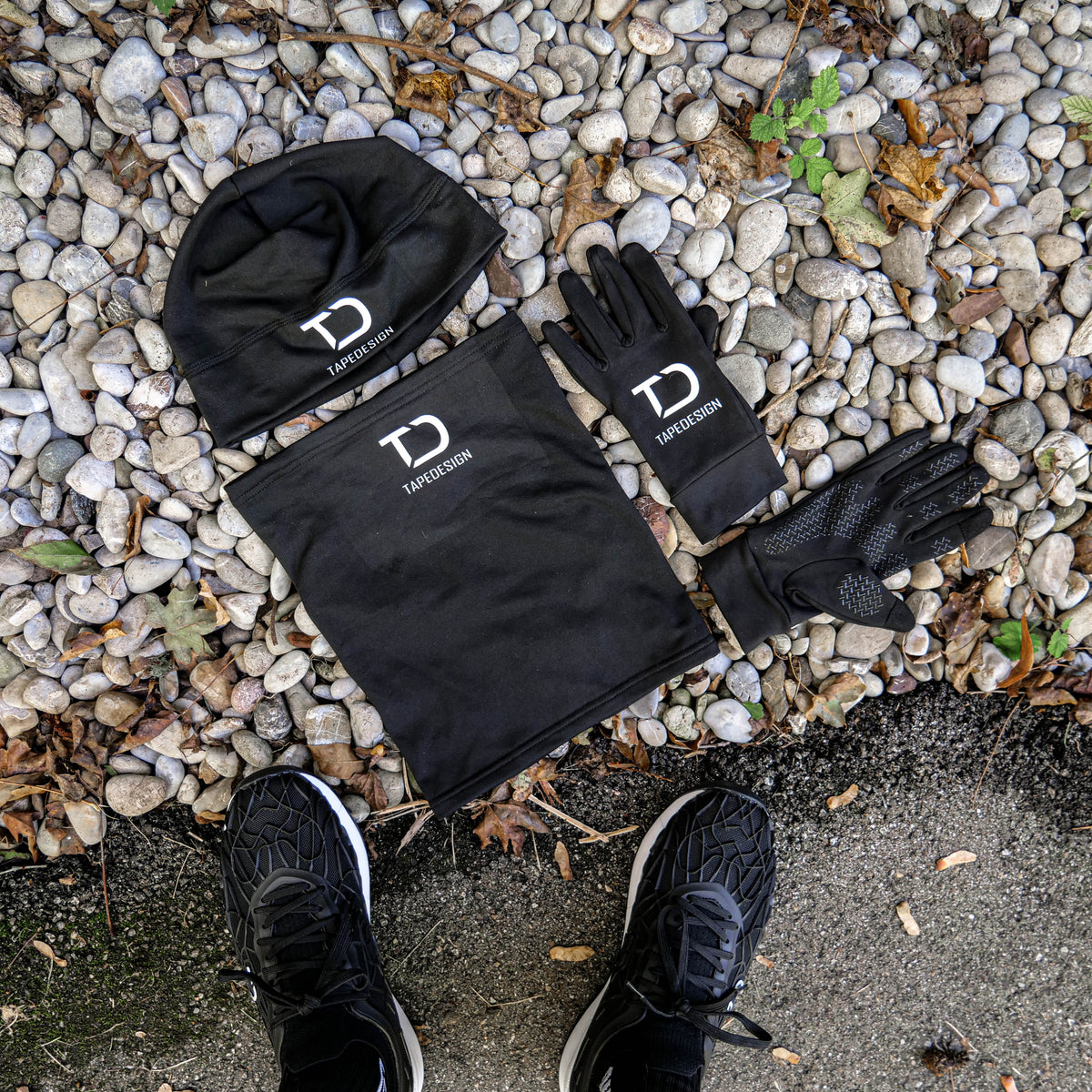 Lieferumfang des schwarzen Tapedesign Wintersets: Beanie, Halswärmer und ein Paar Handschuhe auf Kies